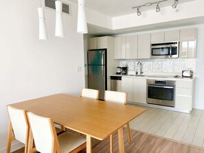 A modern kitchen with white cabinets, stainless steel appliances, and a wooden dining table with chairs. - 1BR Balcony · Bay View · Beachwalk Resort (Hallandale Beach)
