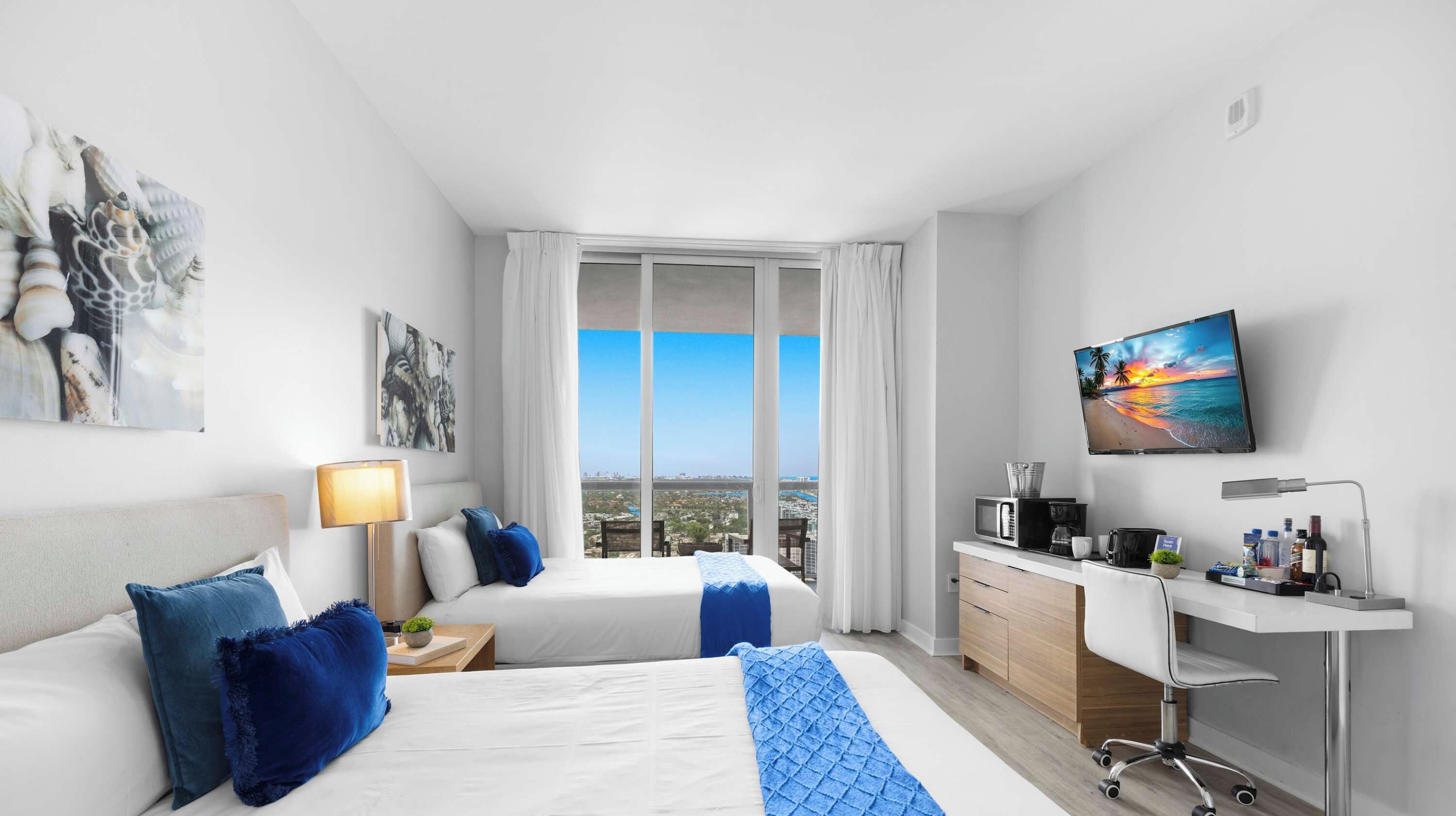 Une chambre d’hôtel équipée d’un lit, d’un bureau, d’une télévision et d’une grande fenêtre donnant sur la ville.