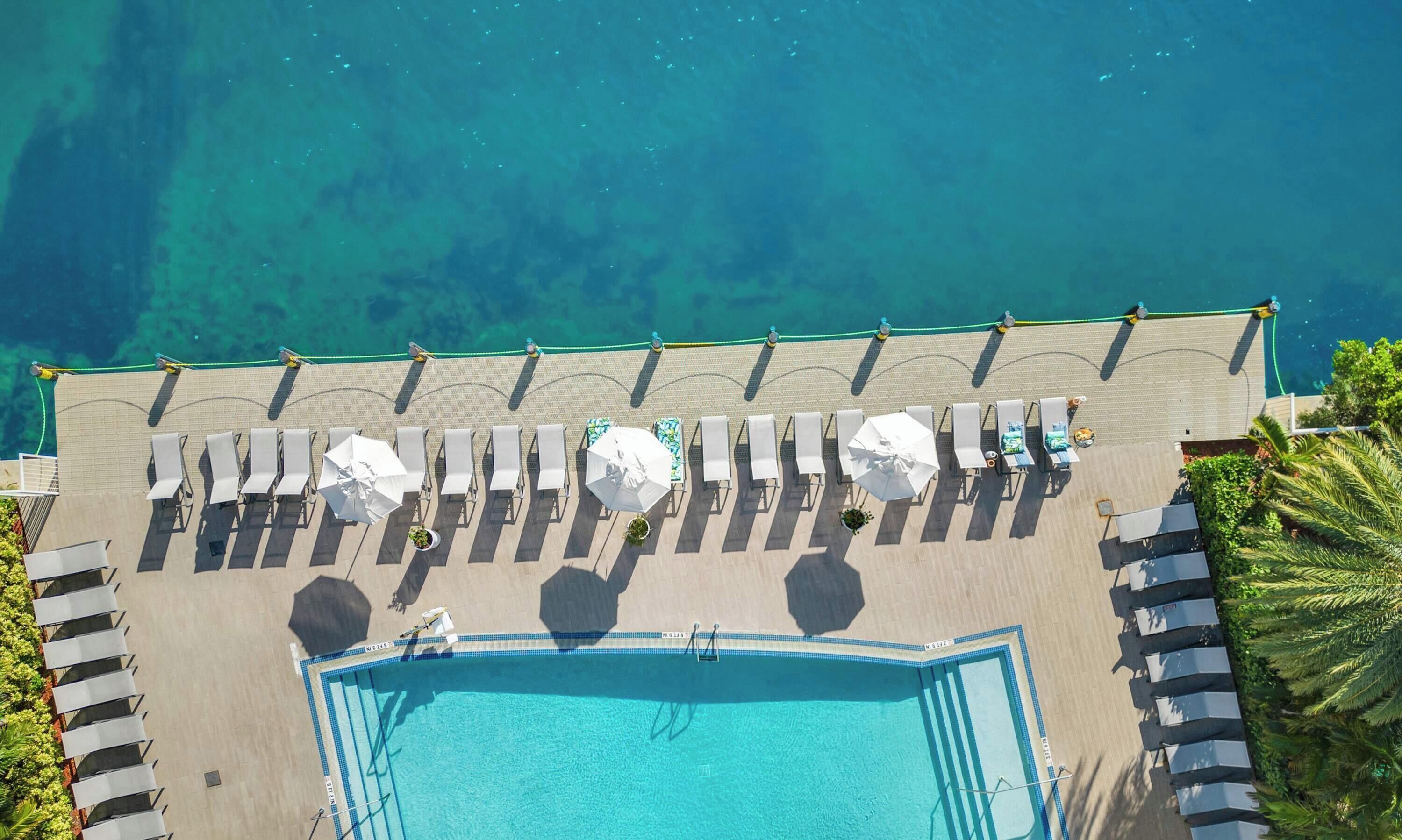 Vue aérienne d’un espace piscine avec des chaises longues et des parasols, entouré d’une piscine à l’eau cristalline et d’une plage de sable.