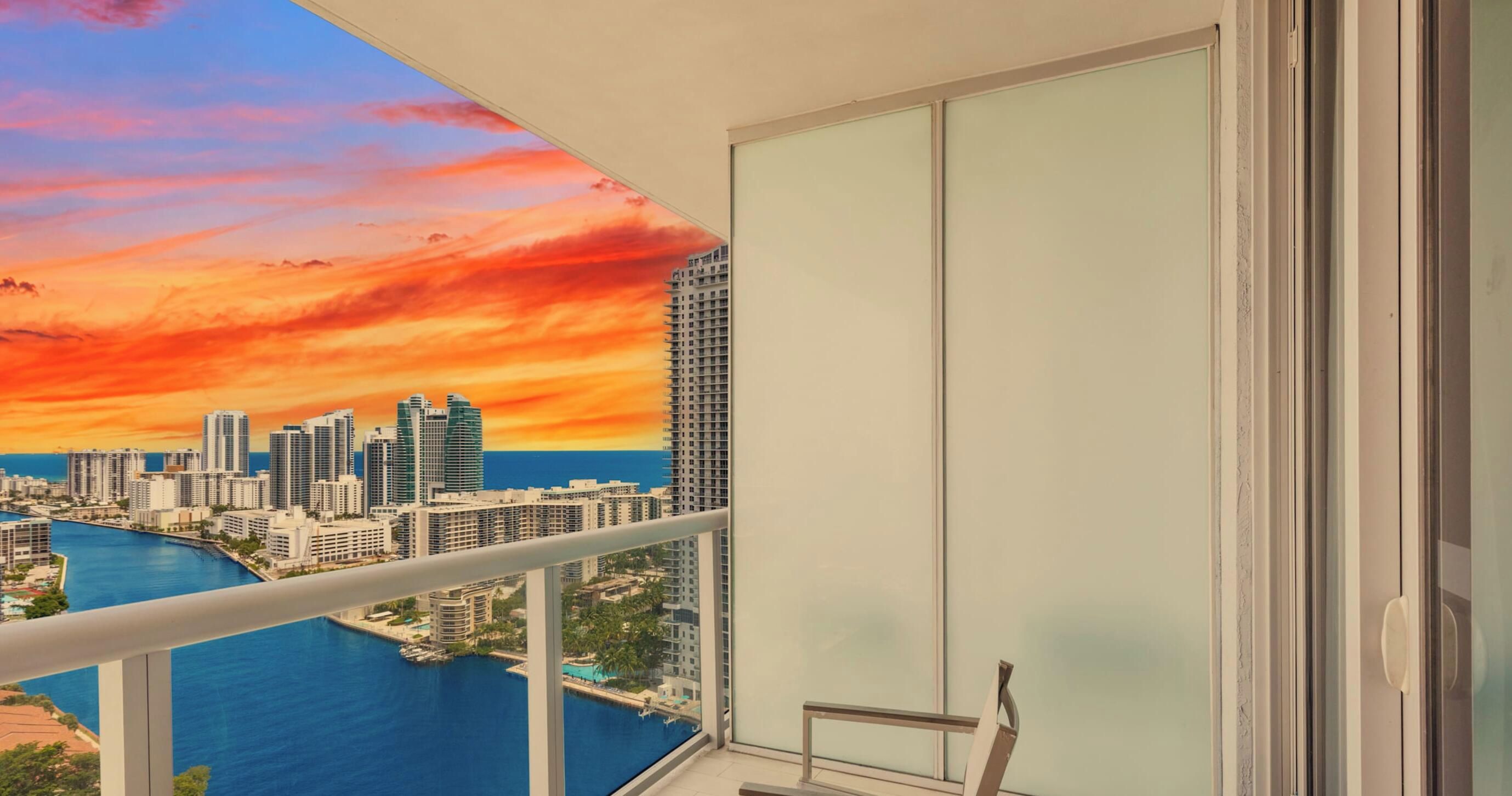 Ein Balkon mit Blick auf die Stadtlandschaft im Sonnenuntergang, eine Glastür und ein Geländer.