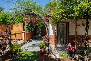 Un jardí amb una pèrgola de fusta, plantes en testos i un camí que condueix a un edifici amb una porta decorada.