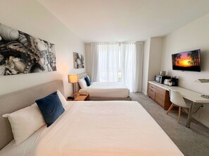A hotel room with two beds, a desk, a television, and artwork on the walls. - Breathtaking Bay View · 2BR · Beachwalk (Hallandale Beach)