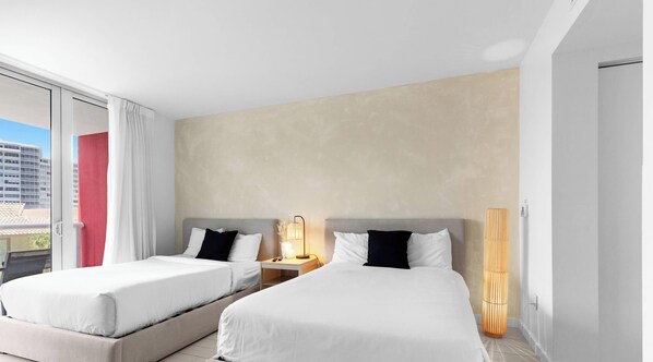 Quarto de hotel com duas camas, uma janela grande com cortinas, abajur e um candeeiro de chão alto e cilíndrico.