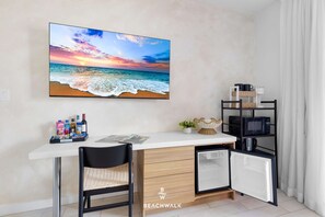 Quarto compacto com uma escrivaninha branca, uma cadeira, um pequeno frigorífico, um micro-ondas e uma estante com utensílios de cozinha. Um quadro grande com uma paisagem de praia está na parede.