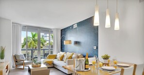 A modern living room with a sofa, dining table, and a large window with a view of palm trees. - Bright 1BR  Waterway View & Resort Lifestyle (Hallandale Beach)