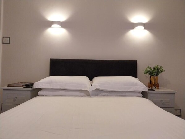 Una cama bien hecha con sábanas blancas, dos mesitas de noche con lámparas, y una pequeña planta en maceta sobre una de ellas.