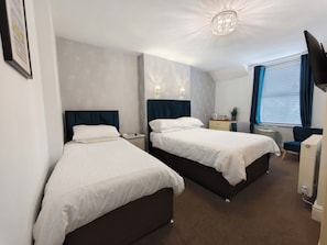A hotel room with two beds, a desk, a chair, a TV, and a window with blinds. - The Rosedene (Llandudno)