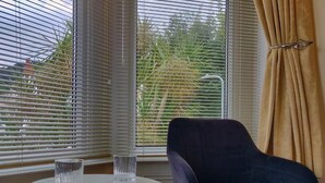 A room with a round table, two chairs, a window with blinds, and a curtain. - The Rosedene (Llandudno)