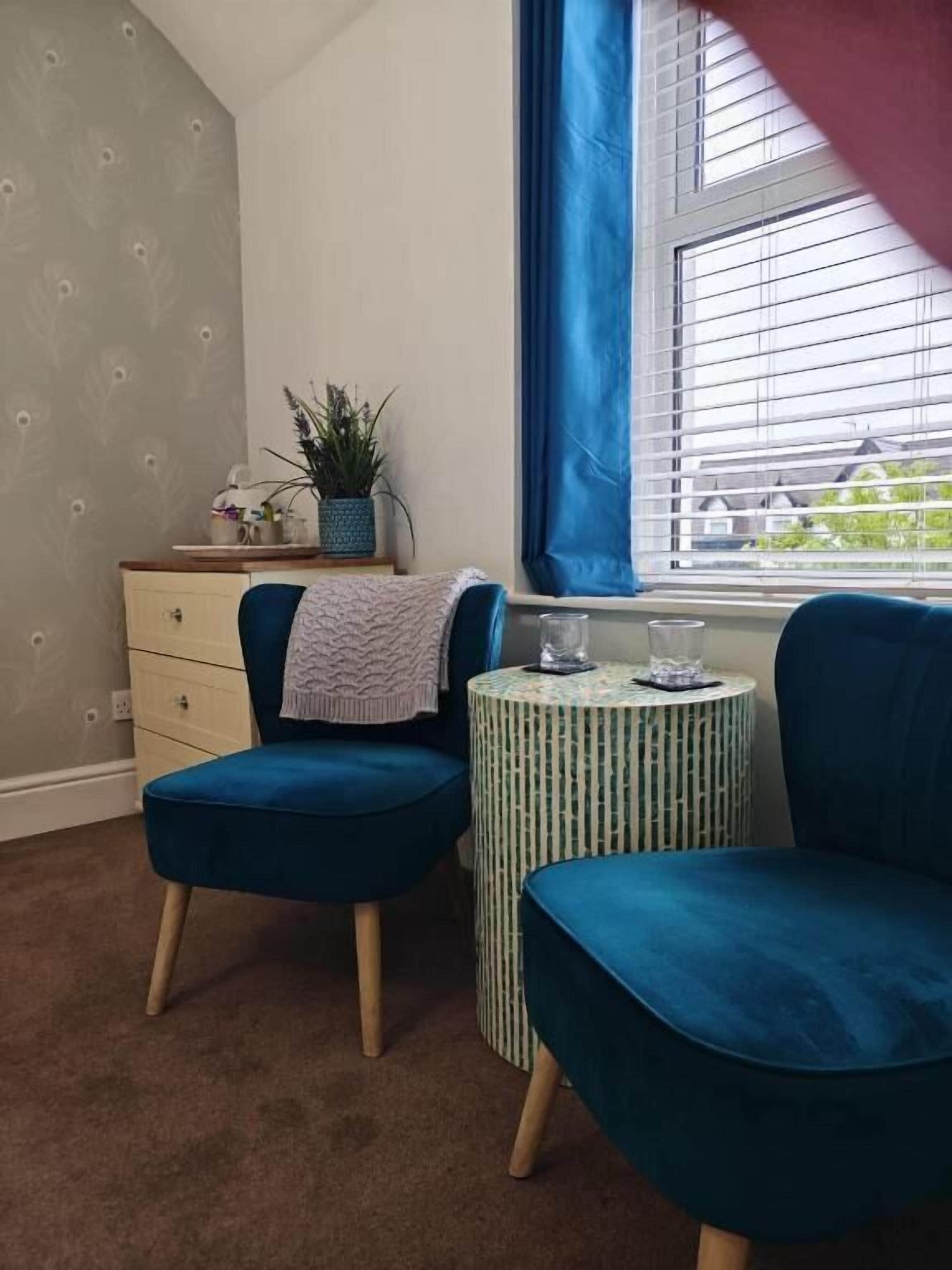 A room with two blue chairs, a small round table, a dresser, a potted plant, and a window with blinds.