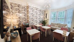 A dining area with a chandelier, a sofa, and a bay window with blinds. - The Rosedene (Llandudno)