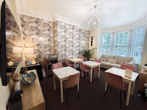 A dining area with a chandelier, a sofa, and a bay window with blinds. - The Rosedene (Llandudno)