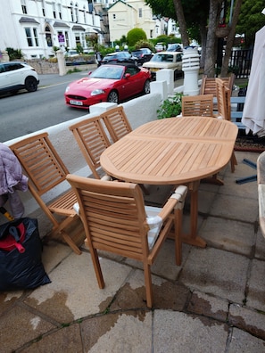 Juego de mesa y sillas de madera al aire libre sobre una acera, con una calle y coches estacionados al fondo.
