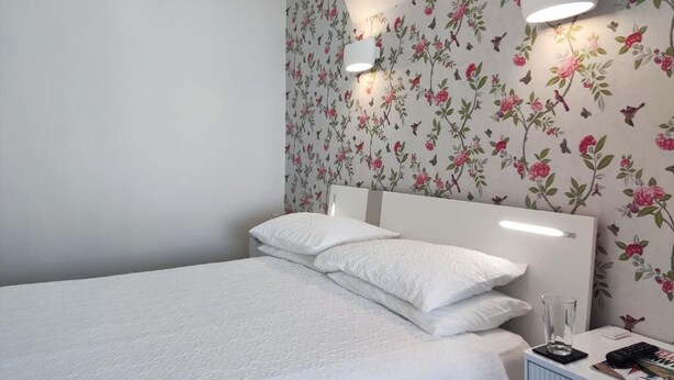 A bedroom with a bed, pillows, a bedside table with a lamp, and a floral wallpaper. - The Rosedene (Llandudno)