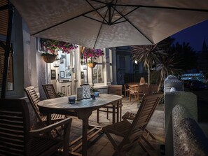 An outdoor patio area with a round table and chairs, a hanging planter with flowers, and a palm tree. - The Rosedene (Llandudno)