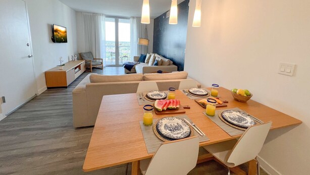 A modern living room with a dining area, featuring a wooden table set for breakfast, a sofa, and a television mounted on the wall. - 1BR with Stunning Intracoastal View, Private Balcony & Full Kitchen (Hallandale Beach)
