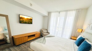 A hotel room with a large bed, a TV mounted on the wall, a wooden TV stand, a chair, and a window with curtains. - 1BR with Stunning Intracoastal View, Private Balcony & Full Kitchen (Hallandale Beach)