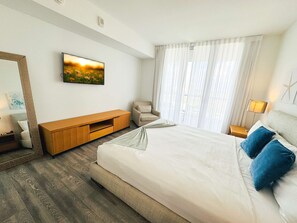 A hotel room with a large bed, a TV mounted on the wall, a wooden TV stand, a chair, and a window with curtains. - 1BR with Stunning Intracoastal View, Private Balcony & Full Kitchen (Hallandale Beach)