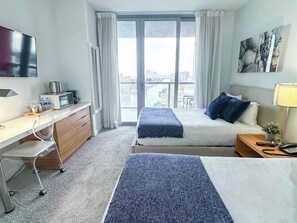 A hotel room with a bed, a desk, a chair, a TV, and a window with a city view. - Beachwalk Studio | Bay & Skyline Vistas (Hallandale Beach)
