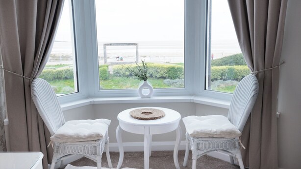A corner room with two white wicker chairs, a small white table, and a view of the sea through large windows. - Bayview Aparthotel by Guestz (Morecambe)