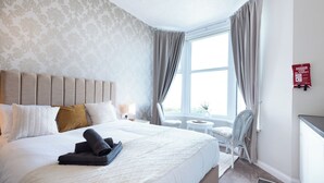 A hotel room with a bed, a window with curtains, a small table, a chair, and a fire blanket sign on the wall. - Bayview Aparthotel by Guestz (Morecambe)