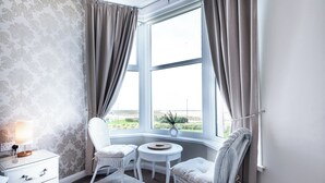 A corner room with a large window, a small round table, two chairs, a nightstand, and a lamp. - Bayview Aparthotel by Guestz (Morecambe)