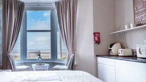 A hotel room with a bed, a small table with a vase, a chair, a view of the beach, a kitchenette, and a fire blanket sign. - Bayview Aparthotel by Guestz (Morecambe)