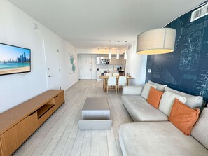 A modern living room with a flat-screen TV, a wooden TV stand, a grey sofa with orange cushions, a dining area with a table and chairs, and a kitchen in the background. - Stunning South Balcony · 2BR Upper Resort Suite (Hallandale Beach)
