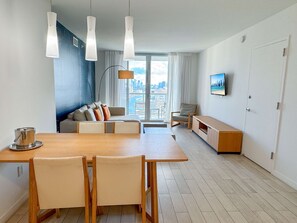 A modern living room with a dining area, a sofa set, a TV mounted on the wall, and a view of the city through large windows. - Stunning South Balcony · 2BR Upper Resort Suite (Hallandale Beach)