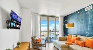 A modern hotel room with a large window, a flat-screen TV, a sofa, a chair, and a map wall. - Top-Floor Bay View · 2BR Beachwalk Resort (Hallandale Beach)