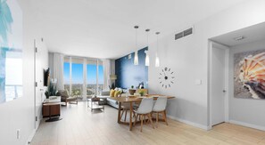 A modern living room with a large window, a dining area with a wooden table, and a kitchen area. - Top-Floor Bay View · 2BR Beachwalk Resort (Hallandale Beach)