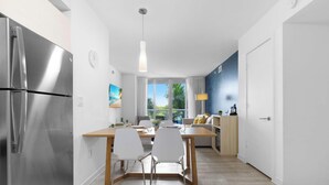 Une cuisine moderne avec un réfrigérateur en acier inoxydable, un coin repas avec une table en bois et des chaises blanches, et une vue sur l’extérieur grâce à de grandes fenêtres.