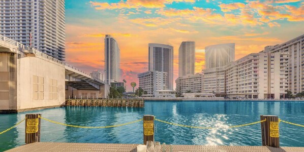 A waterfront view with high-rise buildings, a dock, and a dining setup with a plate of food and drinks. - Elevated Waterway 2BR · Beachwalk Resort (Hallandale Beach)