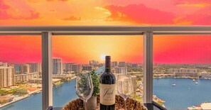 A bottle of wine, two wine glasses, and a woven basket on a balcony with a cityscape view. - Elevated Waterway 2BR · Beachwalk Resort (Hallandale Beach)