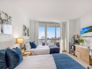 A modern hotel room with a bed, bedside table, lamp, and a view of the city through large windows. - Elevated Waterway 2BR · Beachwalk Resort (Hallandale Beach)