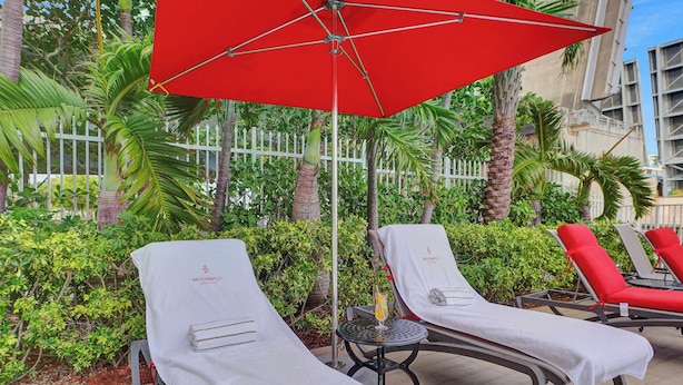 A poolside area with lounge chairs, a red umbrella, and a small table with a drink. - Beach Escape | Beachwalk Resort Studio (Hallandale Beach)