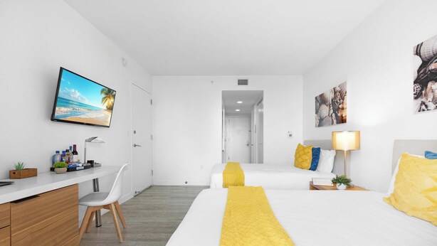 A hotel room with a bed, bedside table, lamp, and a desk with a chair. - Modern Studio in Hallandale | Resort Amenities (Hallandale Beach)