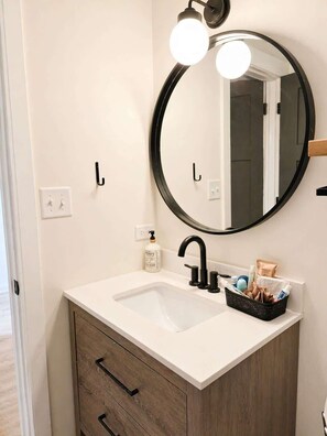 Un baño con un lavabo blanco, grifo negro y un espejo redondo iluminado. Hay dos ganchos negros en la pared, una pequeña botella de jabón y una cesta con artículos de aseo.