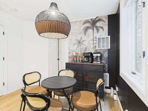 Un comedor con una mesa redonda, cuatro sillas, una lámpara colgante, una cafetera y un armario con botellas.