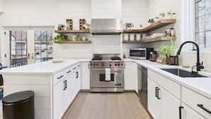 A modern kitchen with white cabinetry, a stainless steel stove, and wooden shelves with plants. - Historic Estate | 10 BR, Sleeps 16+, Game Room (Chicago)
