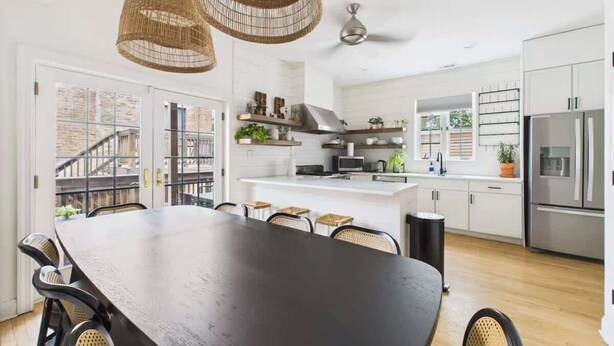 A modern kitchen with white cabinets, a central island, and a dining area with a dark wooden table and wicker chairs. - Historic Estate | 10 BR, Sleeps 16+, Game Room (Chicago)