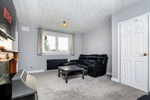 A room with a black leather sofa, a small table, a radiator, a window with curtains, a white door, and a ceiling light. - Spacious 4-Bedroom Home in Torry, Aberdeen – Close to City Centre & Beach (Aberdeen)