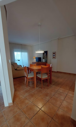 Un área de comedor con una mesa y sillas de madera, una lámpara en el techo, un sofá, un televisor y una ventana con cortinas.