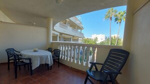 Un balcón con mesa y sillas, barandilla blanca y vista a edificios y palmeras.