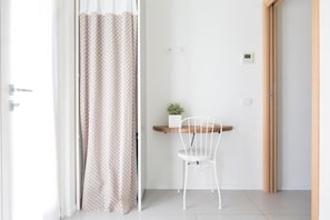 Ein Zimmer mit einer weißen Tür, einem Vorhang mit Punkte-Muster, einem weißen Stuhl, einem kleinen Tisch mit einer Pflanze und einem hölzernen Türrahmen.