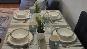 Een eettafel gedekt met borden, kommen, bekers en bestek, met een bloempot en een bank op de achtergrond.