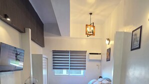 Een slaapkamer met een bed, een grijze bank, een vloerkleed met marmerprint, een staande ventilator en een plafondlamp.
