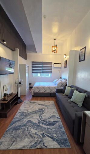 A bedroom with a bed, a grey sofa, a marble-patterned rug, a standing fan, and a ceiling light fixture. - Beautiful studio condo with AC in fabulous Cebu City (Cebu City)