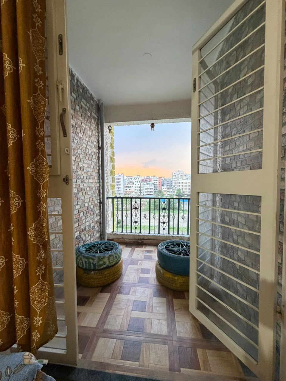 Un balcón con cortinas estampadas, vista panorámica de la ciudad y dos neumáticos grandes como asientos.