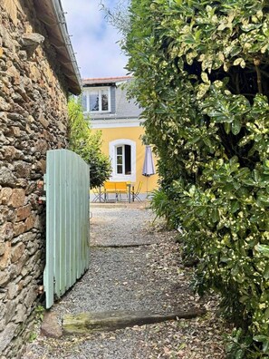 Eine Steinsmauer, ein hölzernes Tor, ein Kiesweg, ein gelbes Gebäude und ein blauer Regenschirm.