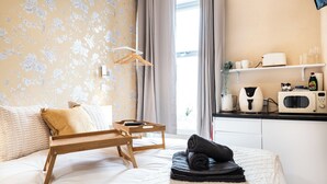 Een hotelkamer met een bed, een houten nachtkastje, een dienblad, een opgerolde handdoek, een raam met gordijnen, een kitchenette met een magnetron, een broodrooster en een plank met kopjes en een fles.
